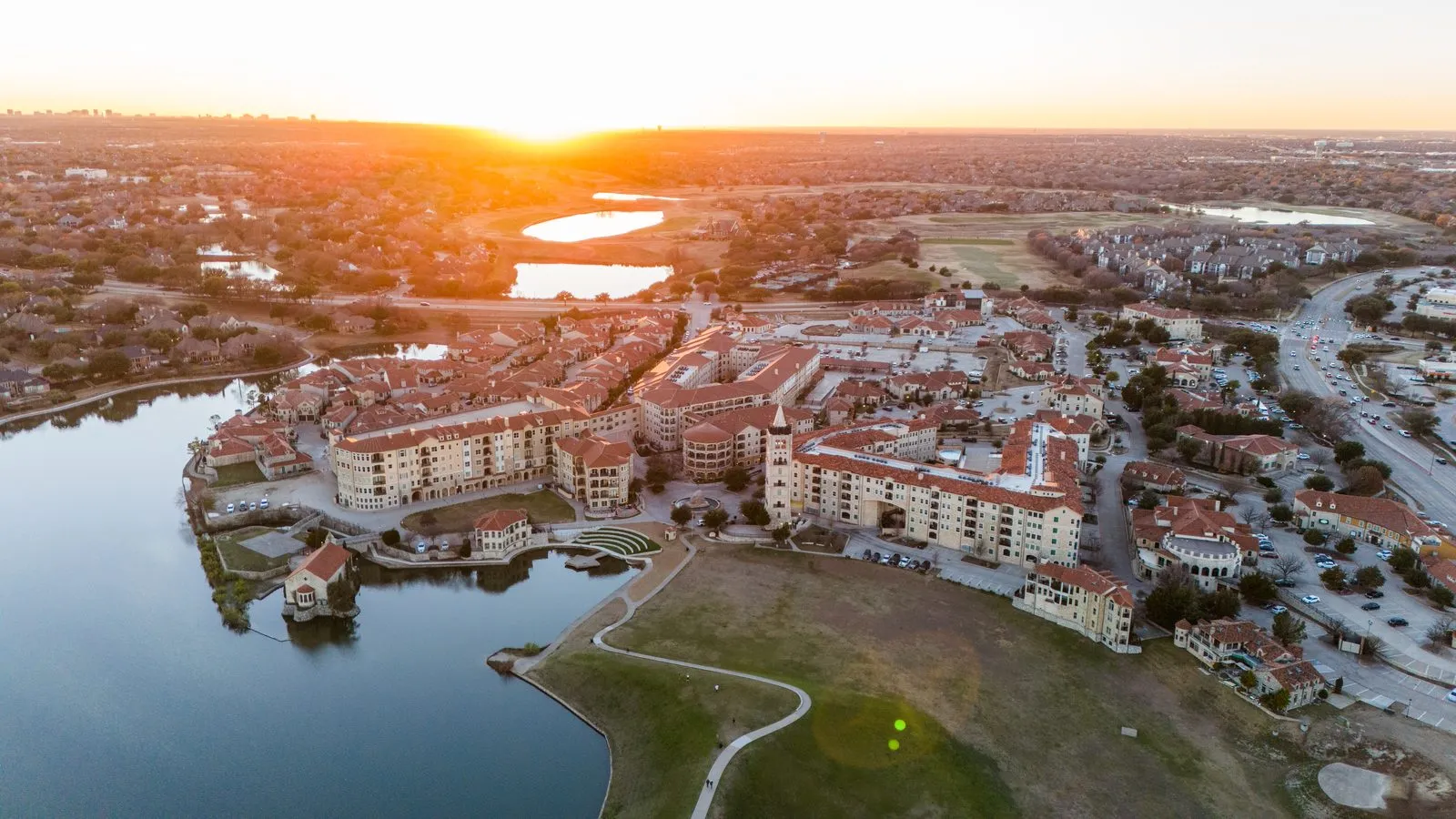 Aerial sunset view of a lakeside Mediterranean-style neighborhood in McKinney, Texas, with red tile rooftops and a marina